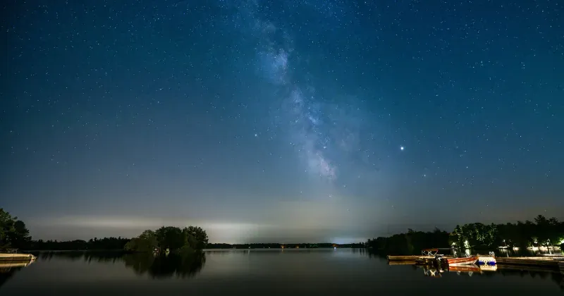 The Milky Way rising over calm dark water, framed by a low tree-lined shore